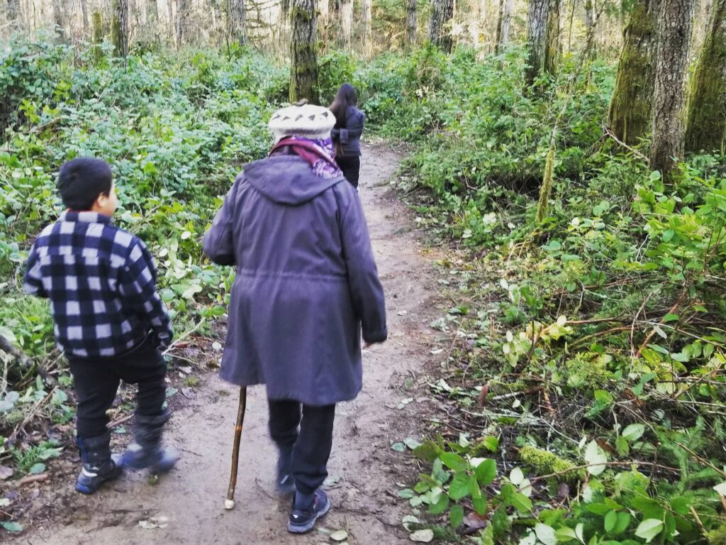 A photo from behind of Indigenous Elder with a walking stick and two Indigenous youth as they walk along Penelakut Trail.