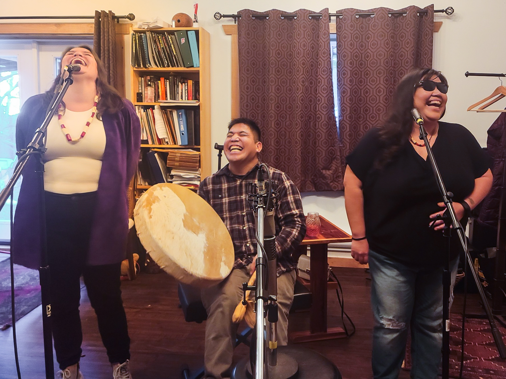 Three First Nations people, two women and a man in the middle are laughing while they record music together. The man is holding a large drum. They are working on Patrick Aleck's new album funded by an FPCC Music Grant.