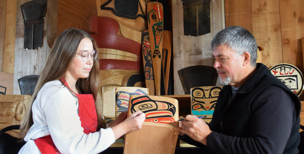 Haida artits Danielle and Christian sit across from each other in a long house painting a bentwood box. Projects like this are supported by the FPCC Arts Strengthening Program