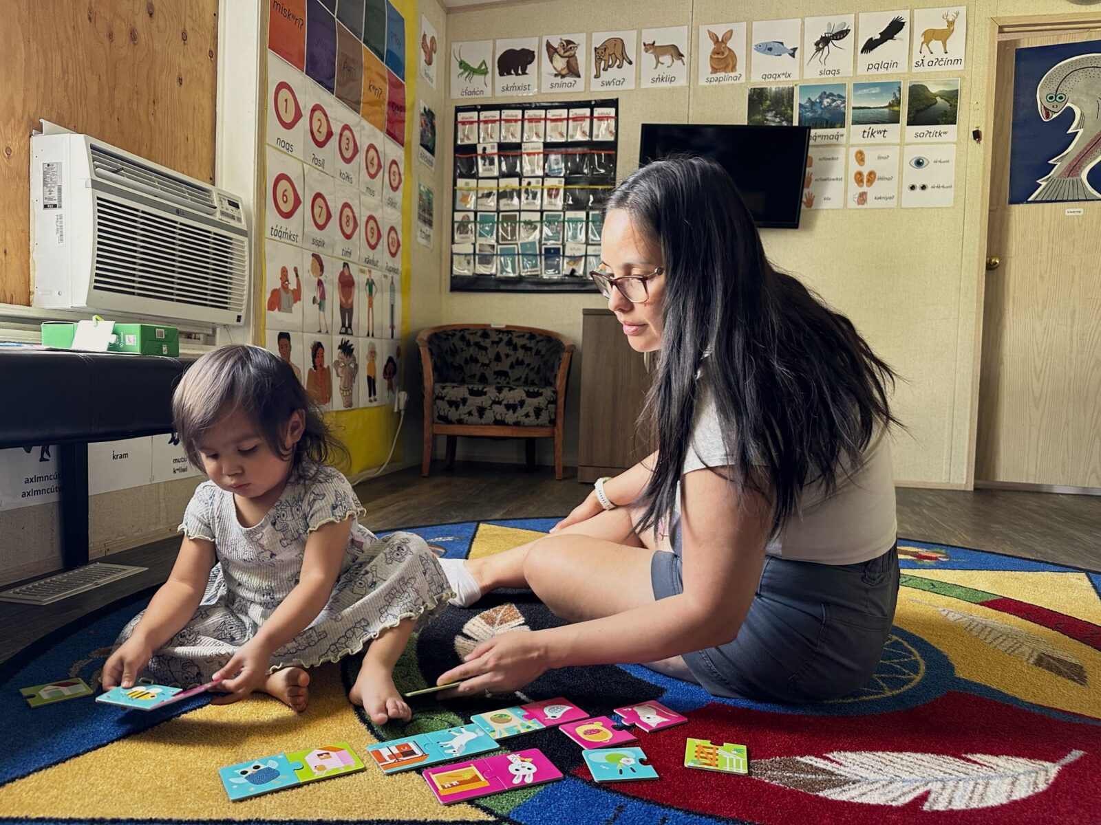 A mother and her child sit on the floor looking at language cards at the nsyilxcen language nest. The image celebrates national Indigenous languages day.