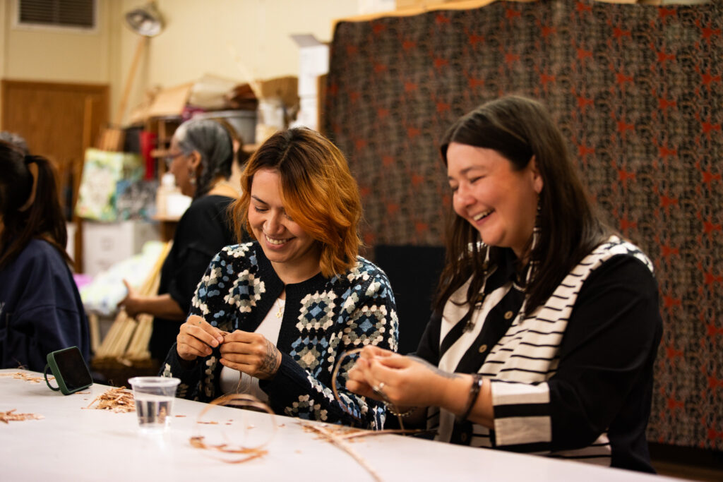 2 people work with cedar bark at a table laughing.