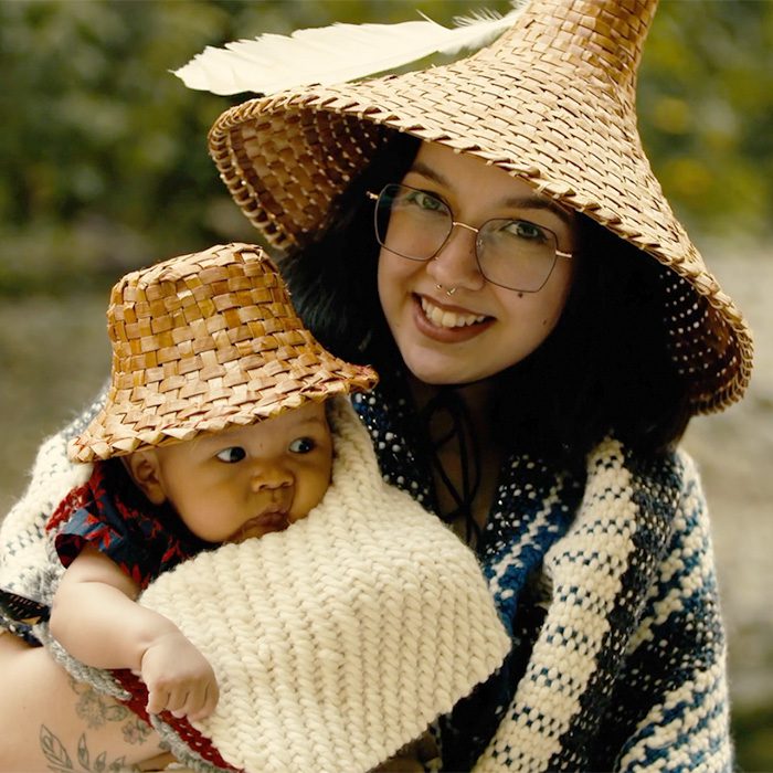 Sarah snuggles her infant son. They are both wrapped in blankets and wearing cedar hats.