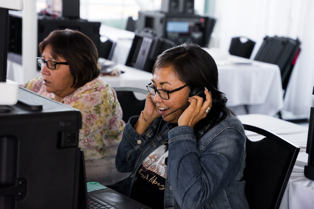 Two people sit in front of electronics at a table. One is smiling and listening to headphones.
