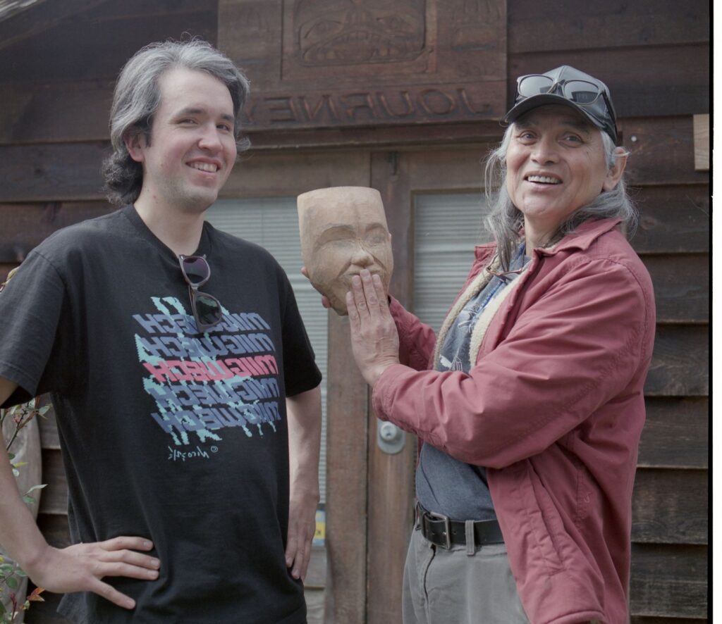 Two First Nations men stand side by side with one holding a carving of a mask depicting a face.