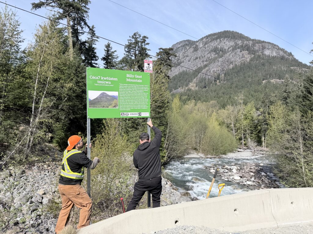 2 people place a sign beside a river at the base of a mountain